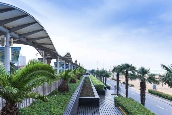 A covered walkway with a curved roof, lined with palm trees and greenery, leading to a road.