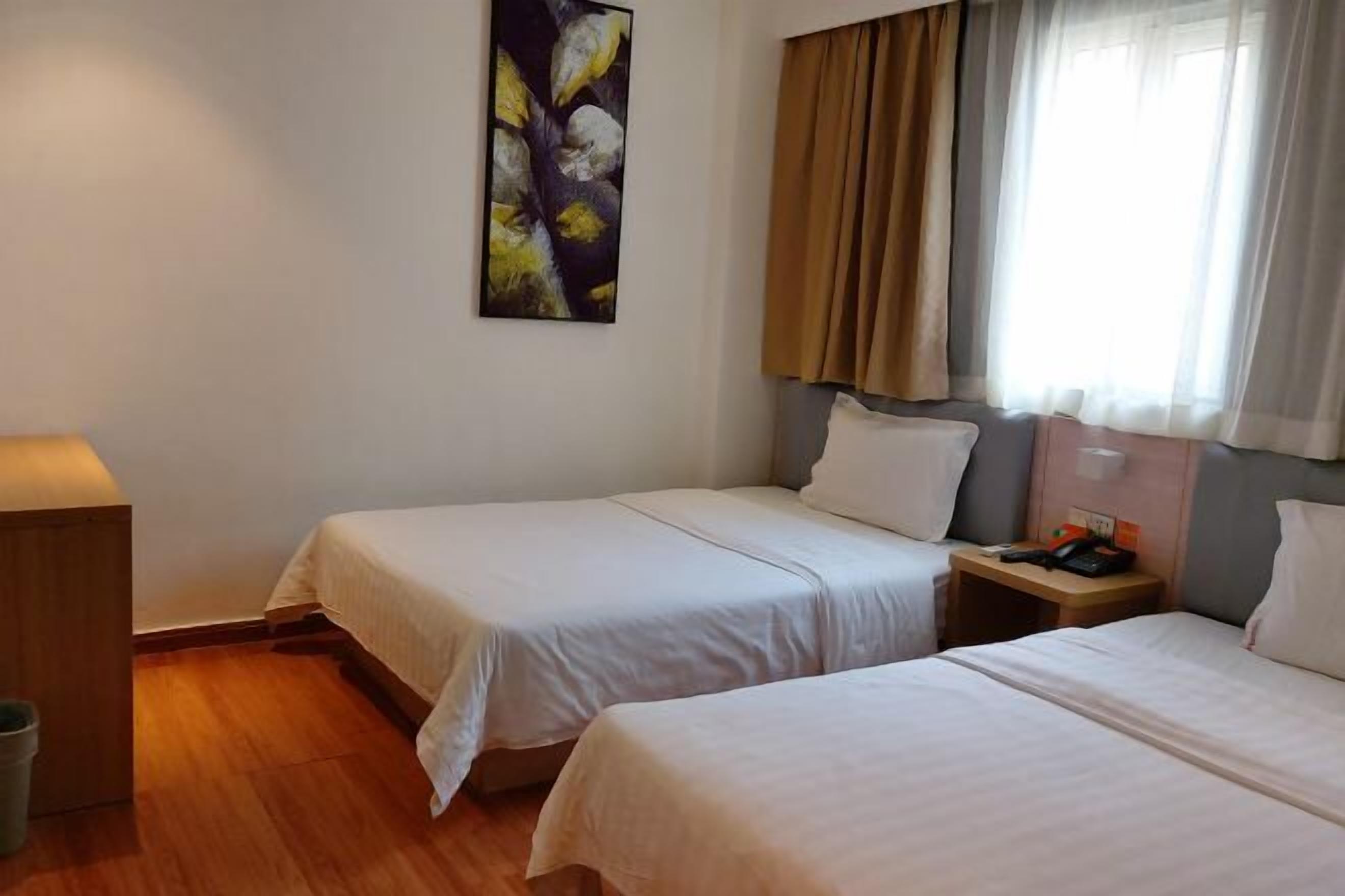 A hotel room with two beds, a nightstand with a phone and a lamp, a painting on the wall, and a window with curtains.