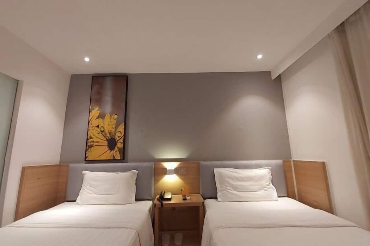 A hotel room with two single beds, a nightstand with a lamp, and a large floral artwork on the wall.