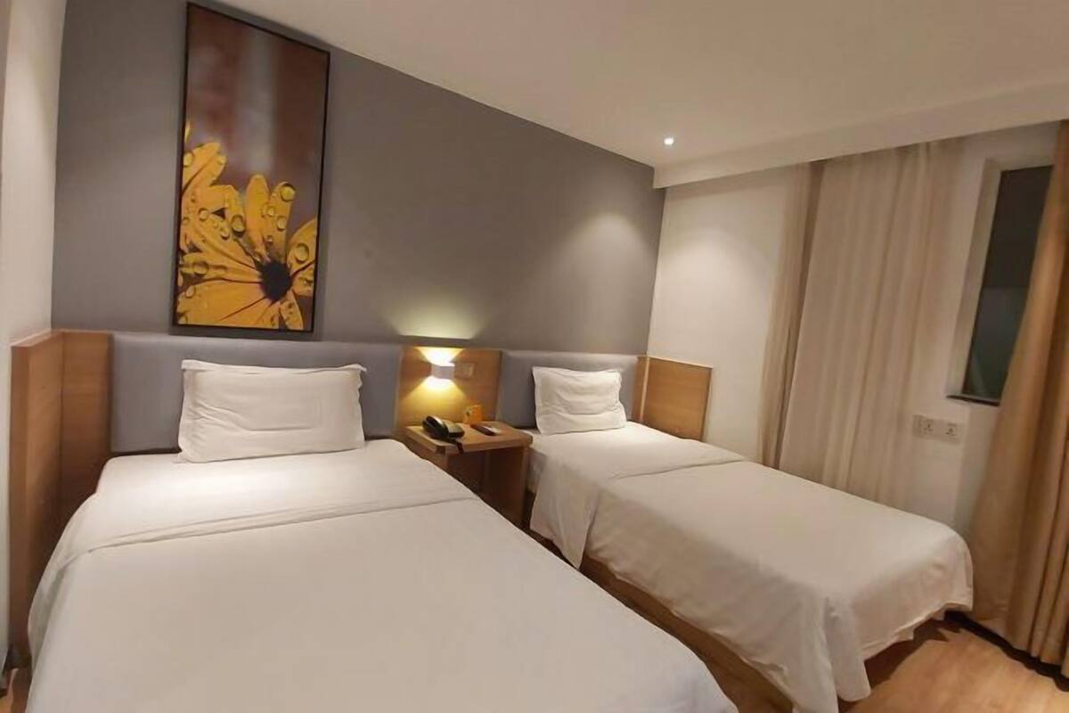 A hotel room with two single beds, a nightstand with a lamp, and a large floral artwork on the wall.