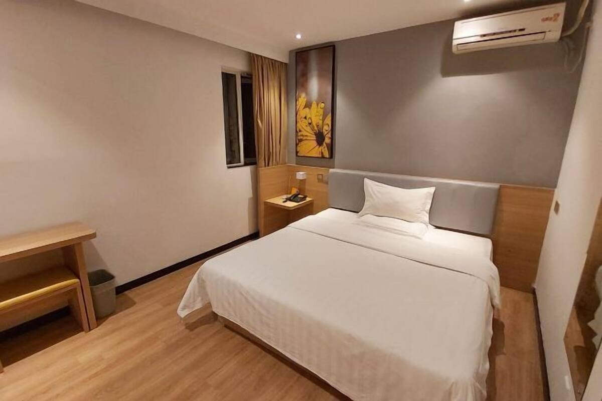 A hotel room with a bed, bedside table, lamp, and an air conditioning unit on the wall.