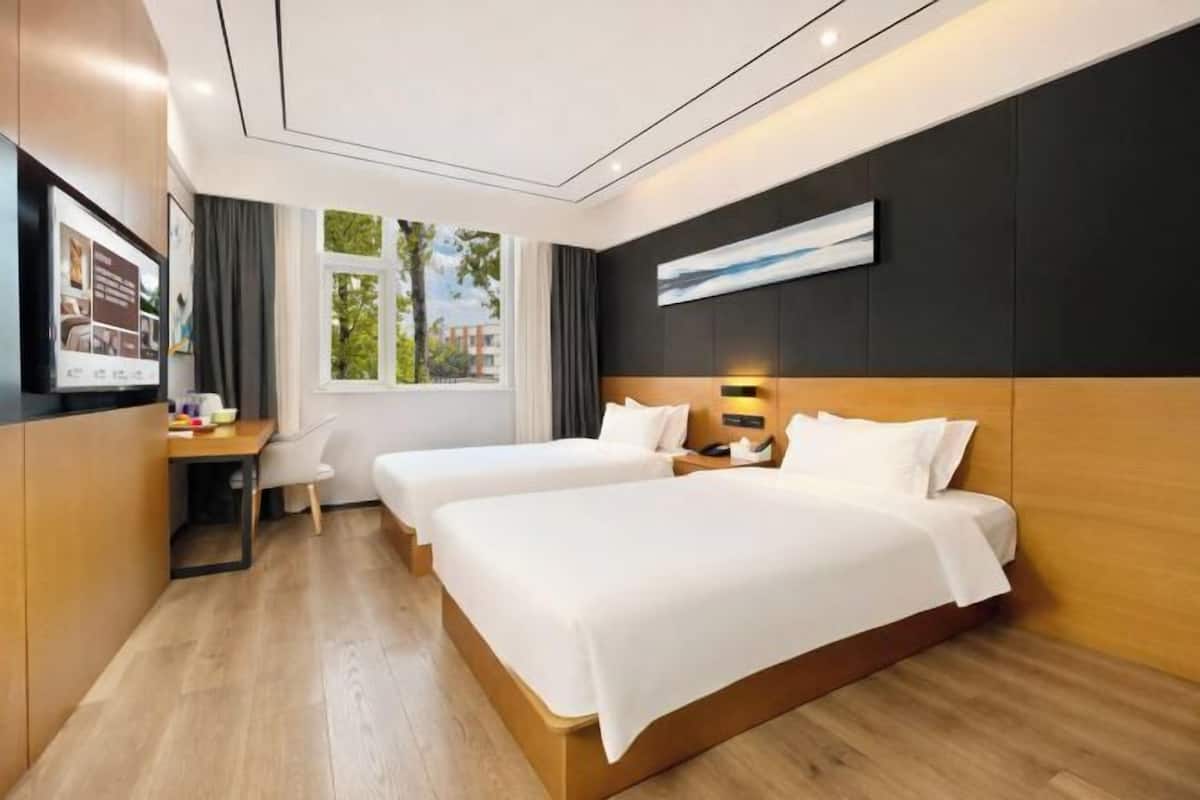 A modern hotel room with two beds, a desk, and a large window offering a view of greenery.