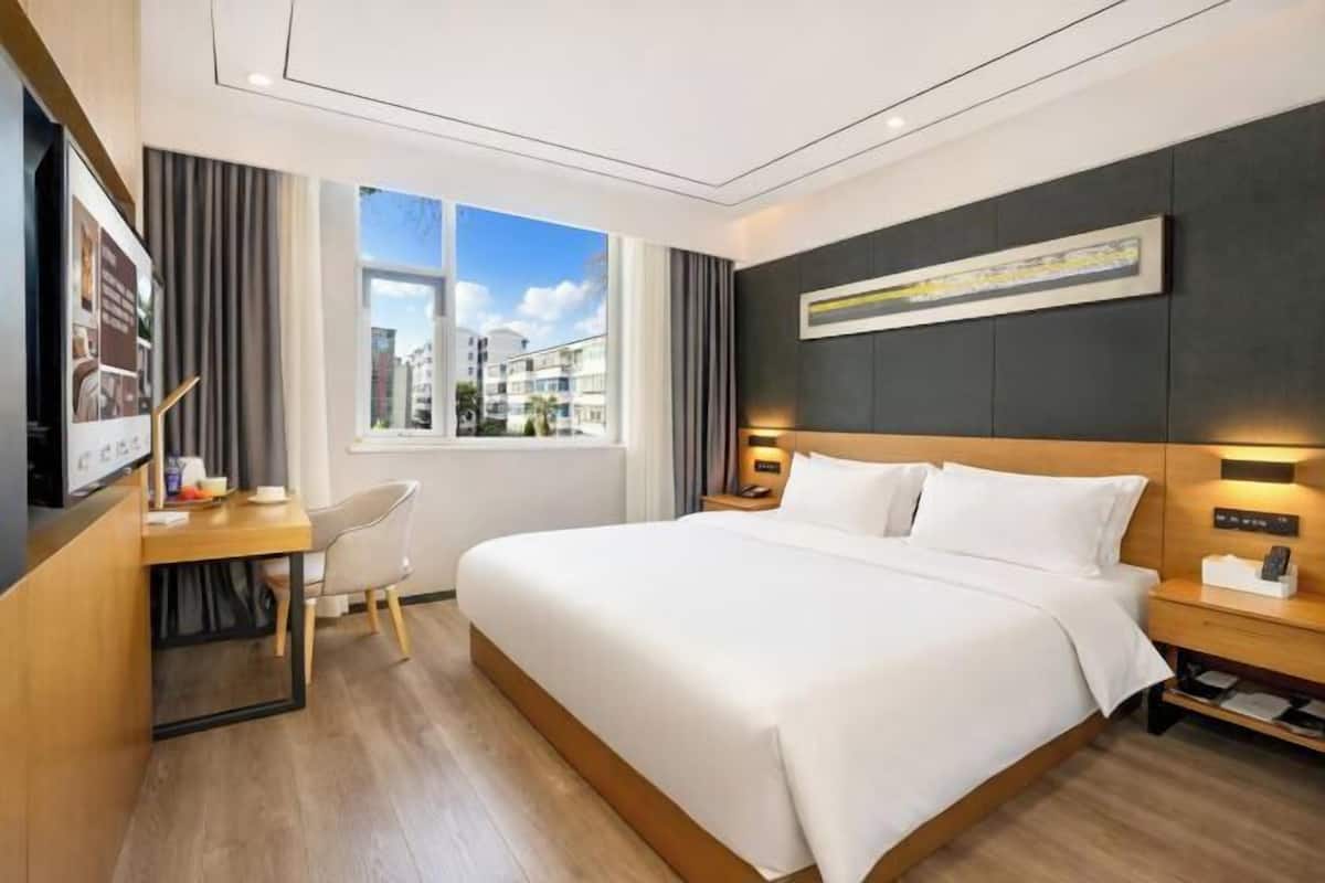 A modern hotel room with a large bed, a desk with a chair, a nightstand, and a window offering a city view.