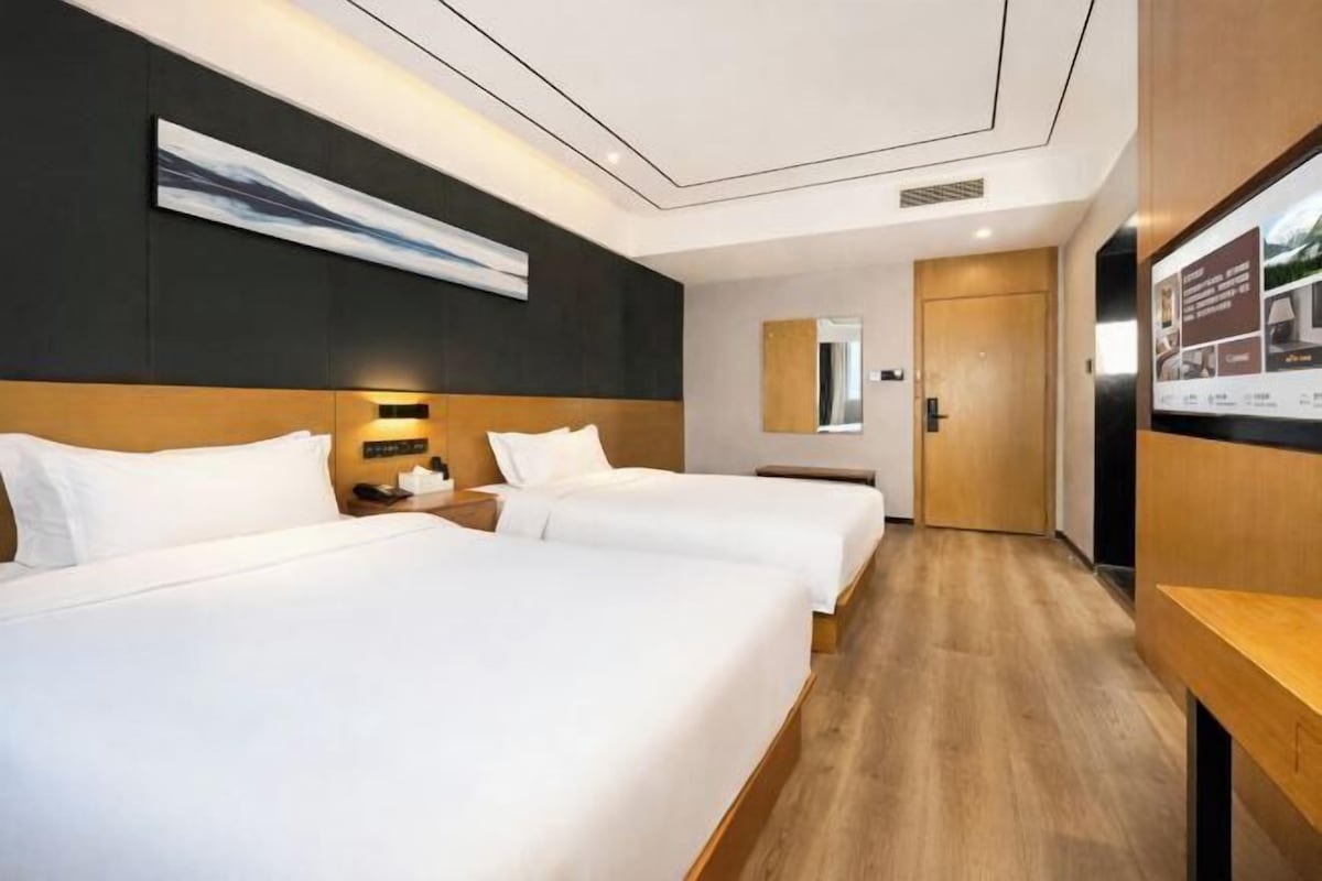 Una habitación de hotel con dos camas, cabecero de madera, mesita de noche, televisor montado en la pared y una puerta que da a otra habitación.