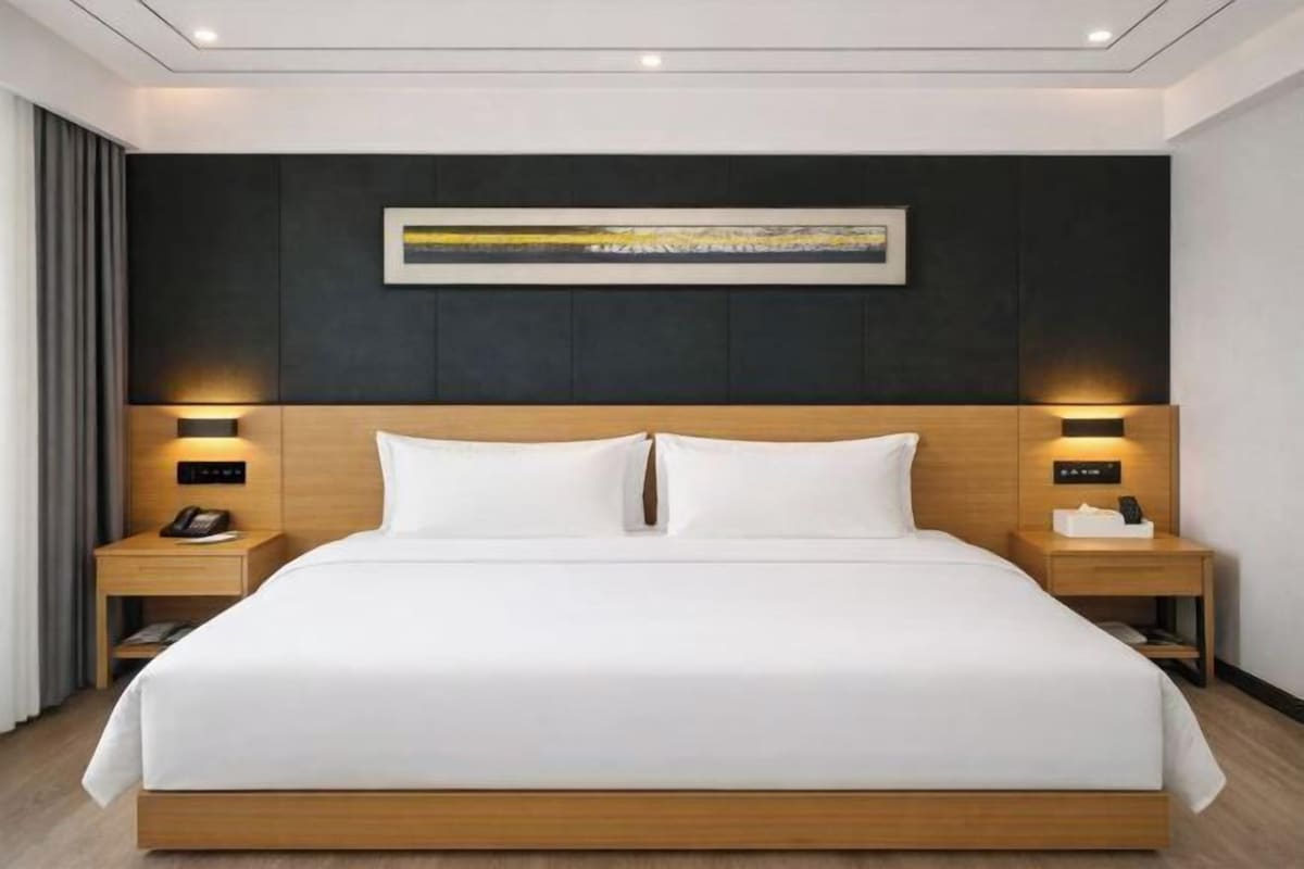 Una habitación de hotel moderna con una cama grande, cabecera de madera, lámparas de noche y una obra enmarcada sobre la cama.