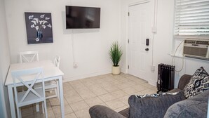 Un salon avec une table blanche, deux chaises blanches, un canapé gris, un téléviseur fixé au mur, une plante en pot et un radiateur.