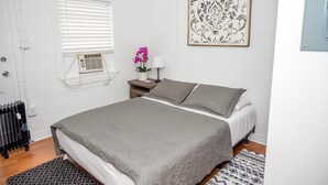 Une chambre à coucher comprenant un lit, une table de chevet, une fenêtre avec des stores, un radiateur, un climatiseur mural, un tableau encadré et un tapis à motifs.