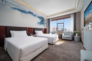 Quarto de hotel com duas camas, área de estar com uma cadeira e uma mesa, uma janela grande com vista para a cidade e um quadro na parede.