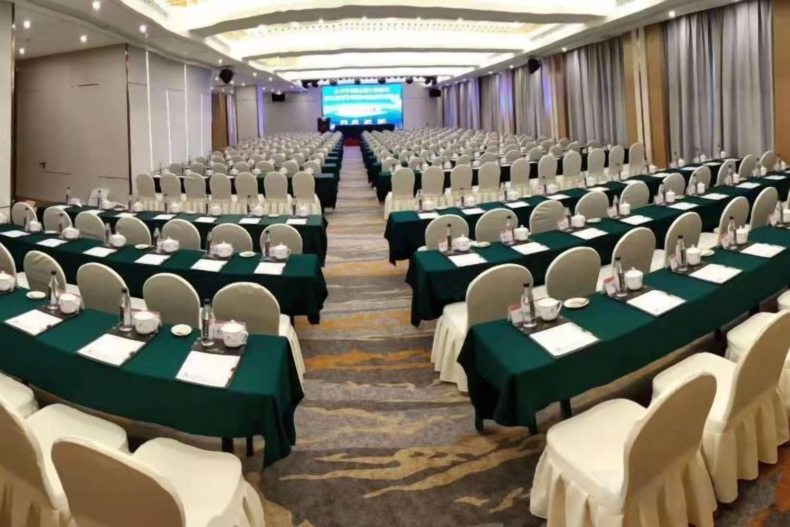 A conference room with rows of tables and chairs, a stage at the front, and a carpeted floor.
