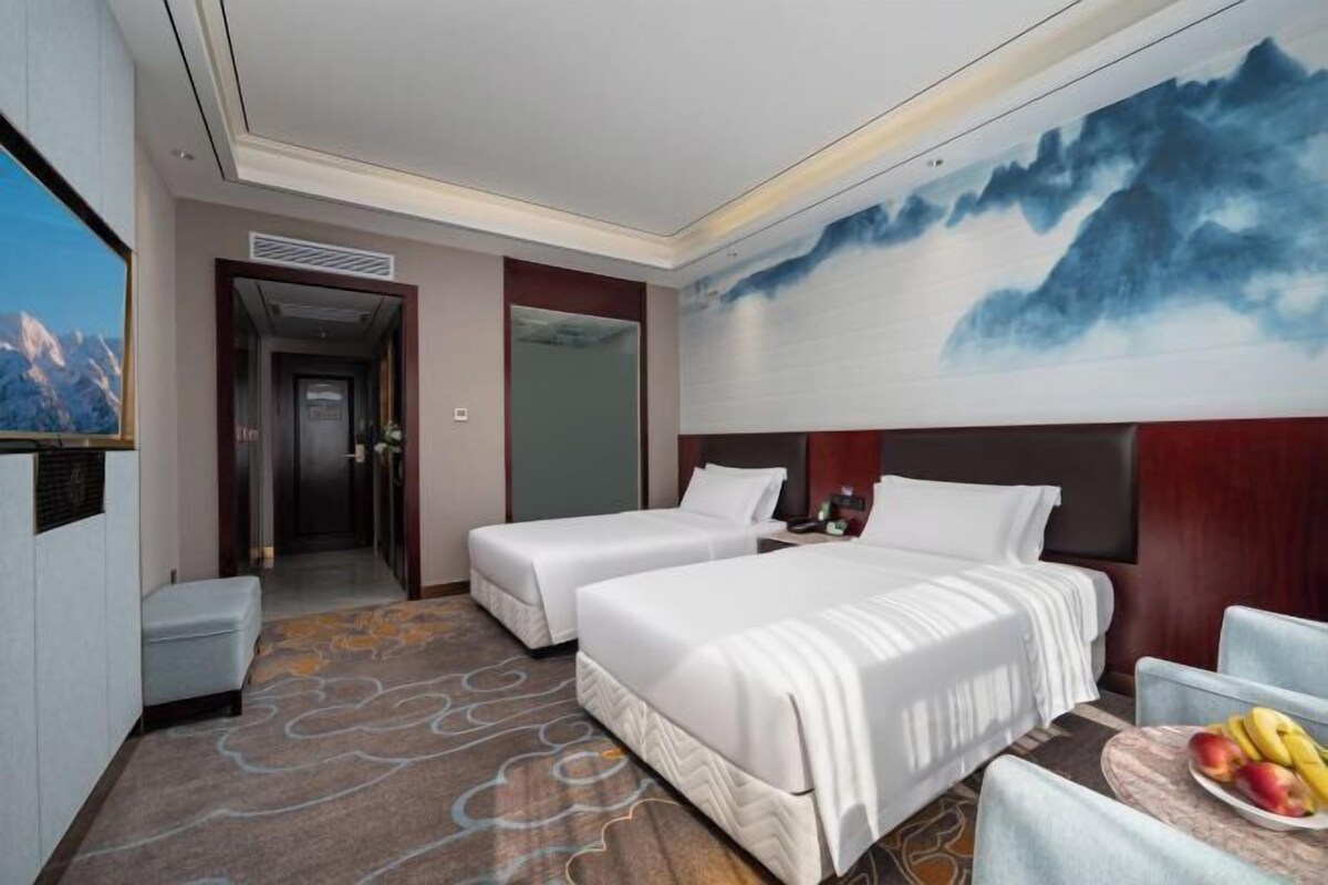 A hotel room with two beds, a small seating area, a painting of mountains, and a view of the mountains through a window.