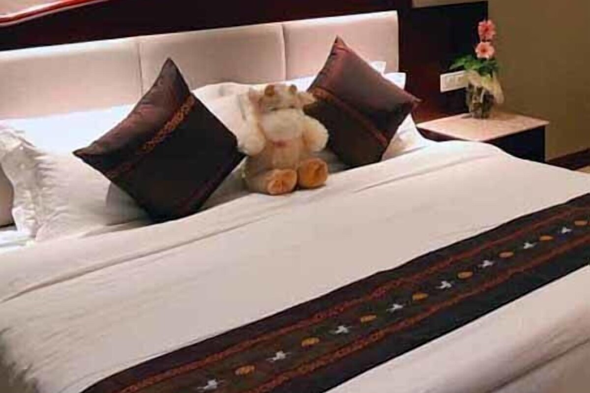 Un lit bien fait, avec une couette blanche, deux oreillers marron foncé et un petit animal en peluche.