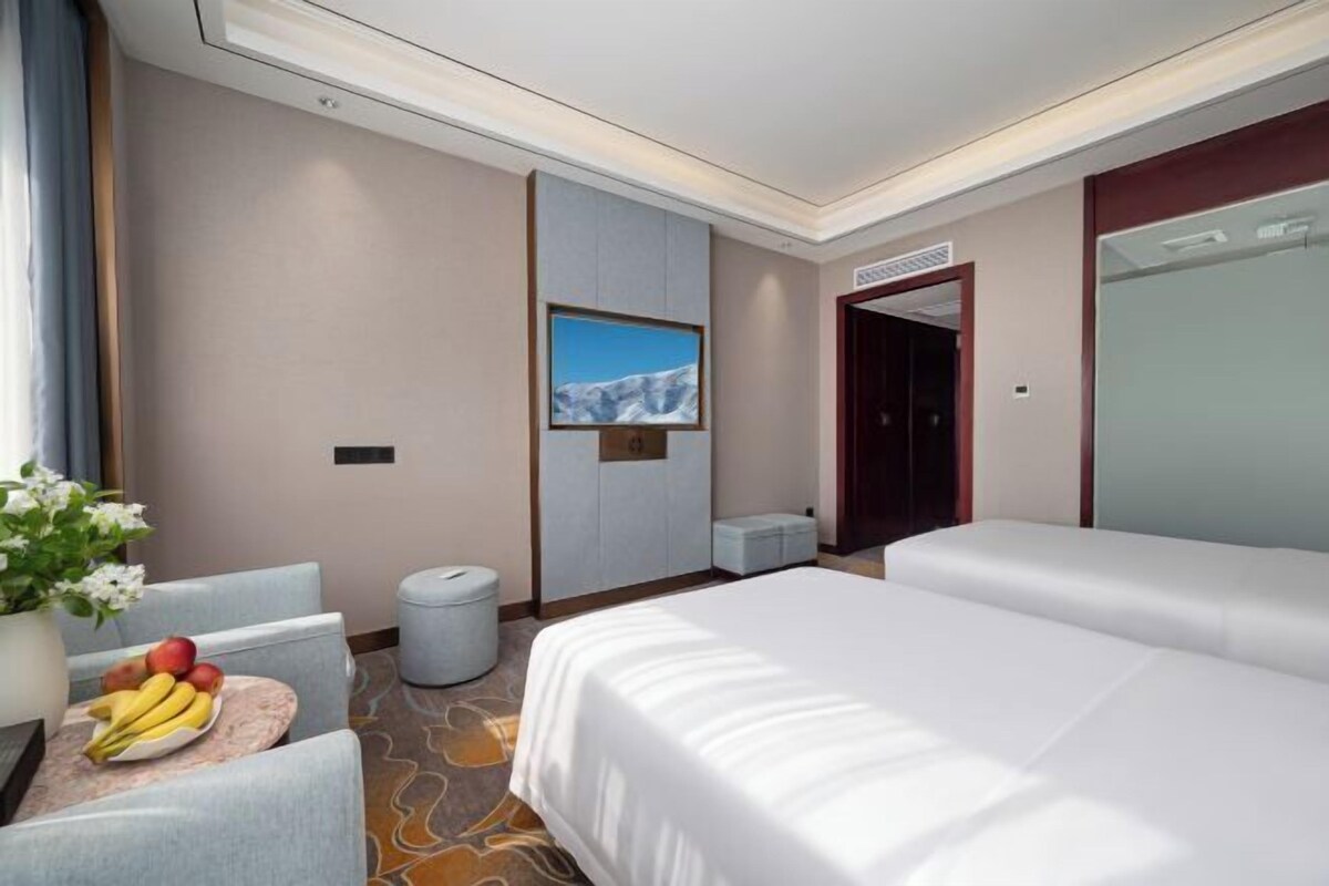Une chambre d’hôtel comprenant un lit, un bureau avec un panier de fruits, une chaise, une petite table, un téléviseur fixé au mur et une porte.