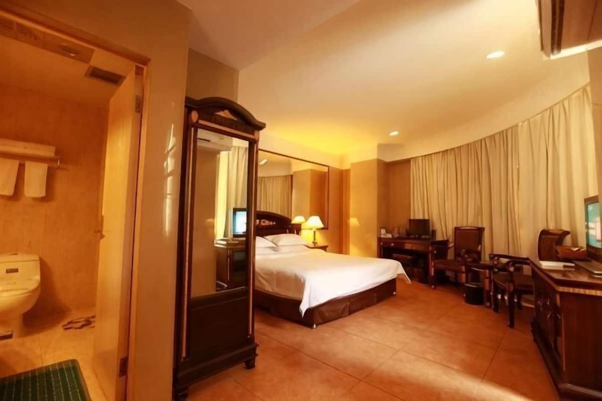 A hotel room with a four-poster bed, a desk with a computer, a chair, a TV, and a bathroom with a toilet and sink.