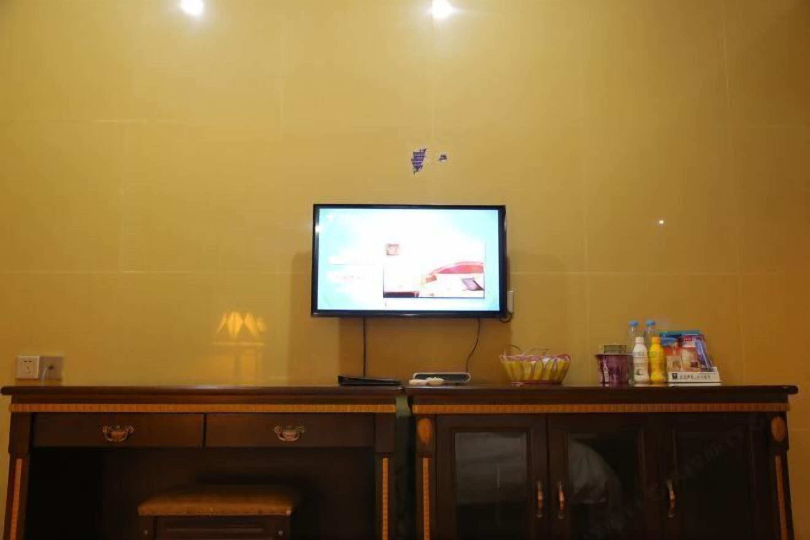 Habitación de hotel con una televisión montada en la pared, un mueble de madera con cajones y una mesita con varios objetos encima.