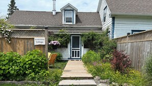 Une maison de campagne dotée d’une terrasse en bois, d’un jardin agrémenté de diverses plantes et fleurs, et d’une pancarte indiquant : « Maison d’été avec cuisine ».