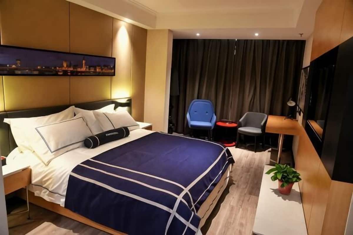 A hotel room with a bed, a desk, a chair, a TV, and a cityscape painting on the wall.