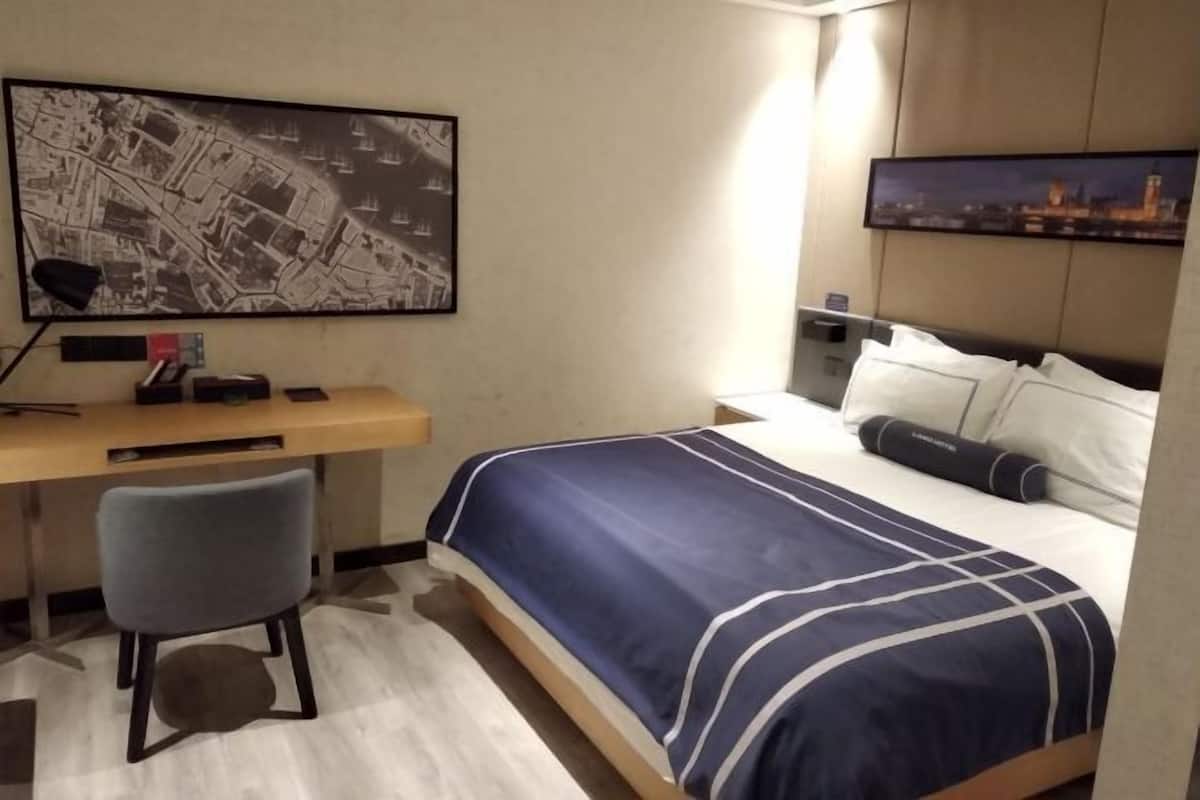 A hotel room with a bed, a desk, a chair, a map on the wall, and a cityscape picture.