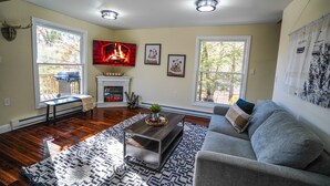 A living room with hardwood floors, a fireplace, a gray sofa, and a large window with a view of trees. - BLVE Cabin w/ Hot Tub,Sauna,Cold Plunge,Game Room Near Bushkill Falls & Shawnee (East Stroudsburg)