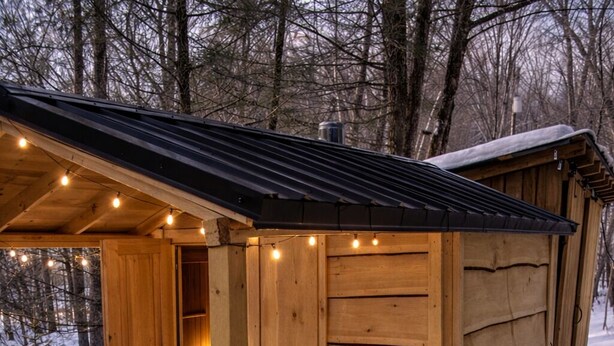 A wooden spa cabin with a hot tub, surrounded by snow, featuring a wooden door, string lights, and a small potted plant.