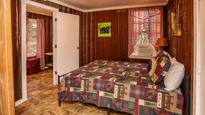 A bedroom with a quilted bedspread, a wooden floor, a red heater, and a window with checkered curtains. - Sasquatch Cabin (Benton)