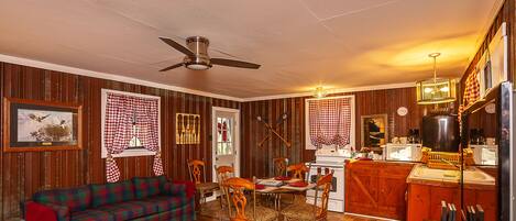A cozy living room with a plaid sofa, wooden dining table, and chairs. A ceiling fan and a chandelier are visible.