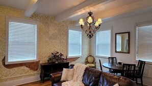 A living room with a brown leather couch, a dining table with chairs, a chandelier, and large windows with blinds. - 502 W Robertson St. Memphis, TX · Converted Historic Jailhouse (Memphis)