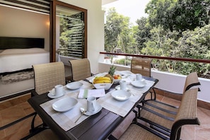 Une table à manger dressée pour deux personnes, avec de l’assiette, des bols et des tasses blancs, donnant sur un jardin luxuriant.