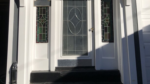 Ein Hauseingang mit einer weißen Tür, zwei Glasfeldern mit geometrischen Mustern und einer kleinen Treppe, die zum Eingang führt.