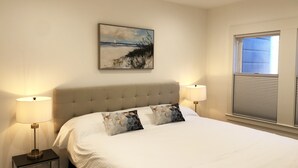 Ein Schlafzimmer mit einem großen Bett, zwei Nachttischlampen, einem Kopfteil, einem Bild an der Wand und einem Fenster mit Jalousien.