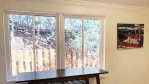 Ein Zimmer mit einem dunklen Holztisch und zwei weißen, gepolsterten Hocker. Ein großes Fenster bietet Blick auf einen Holzzaun und Bäume.