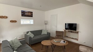 A modern living room with a grey sectional sofa, a round coffee table, a leather armchair, and a flat-screen TV mounted on the wall. - Warm, modern house 50m from the sea (Trébeurden)