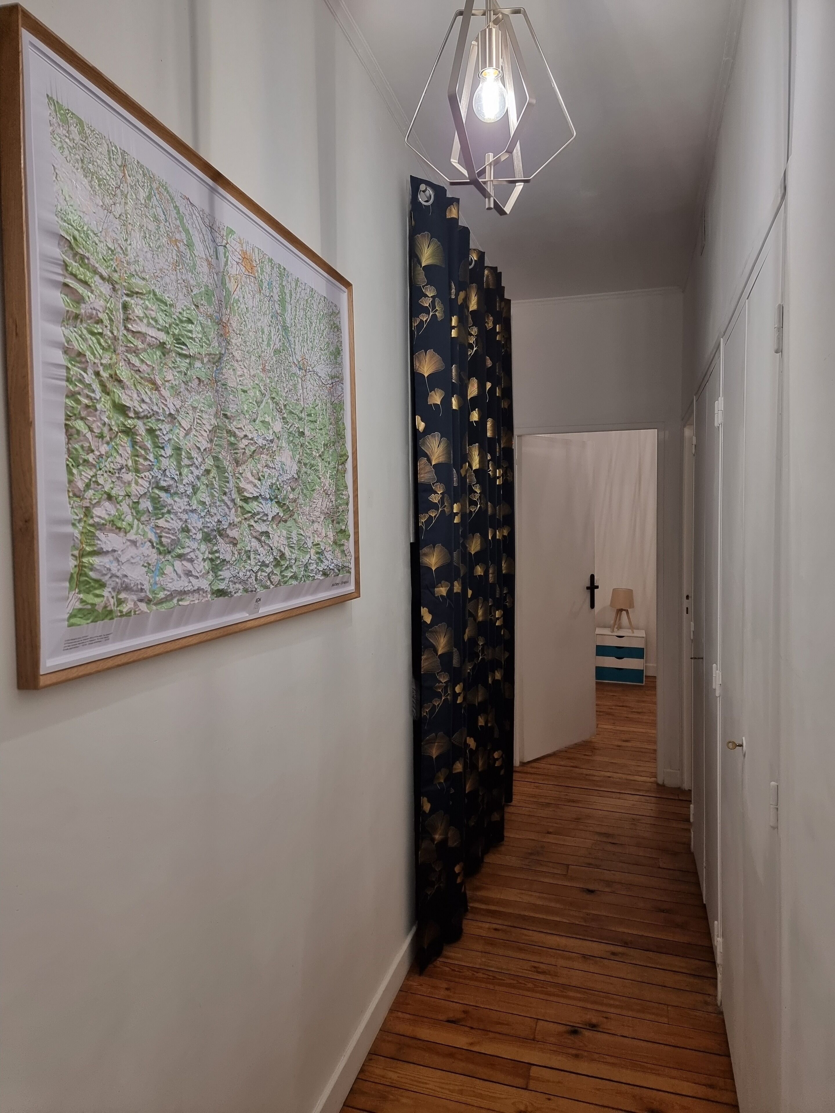 A hallway with a wooden floor, a large map on the wall, a black curtain with a leaf pattern, and a hanging light fixture.