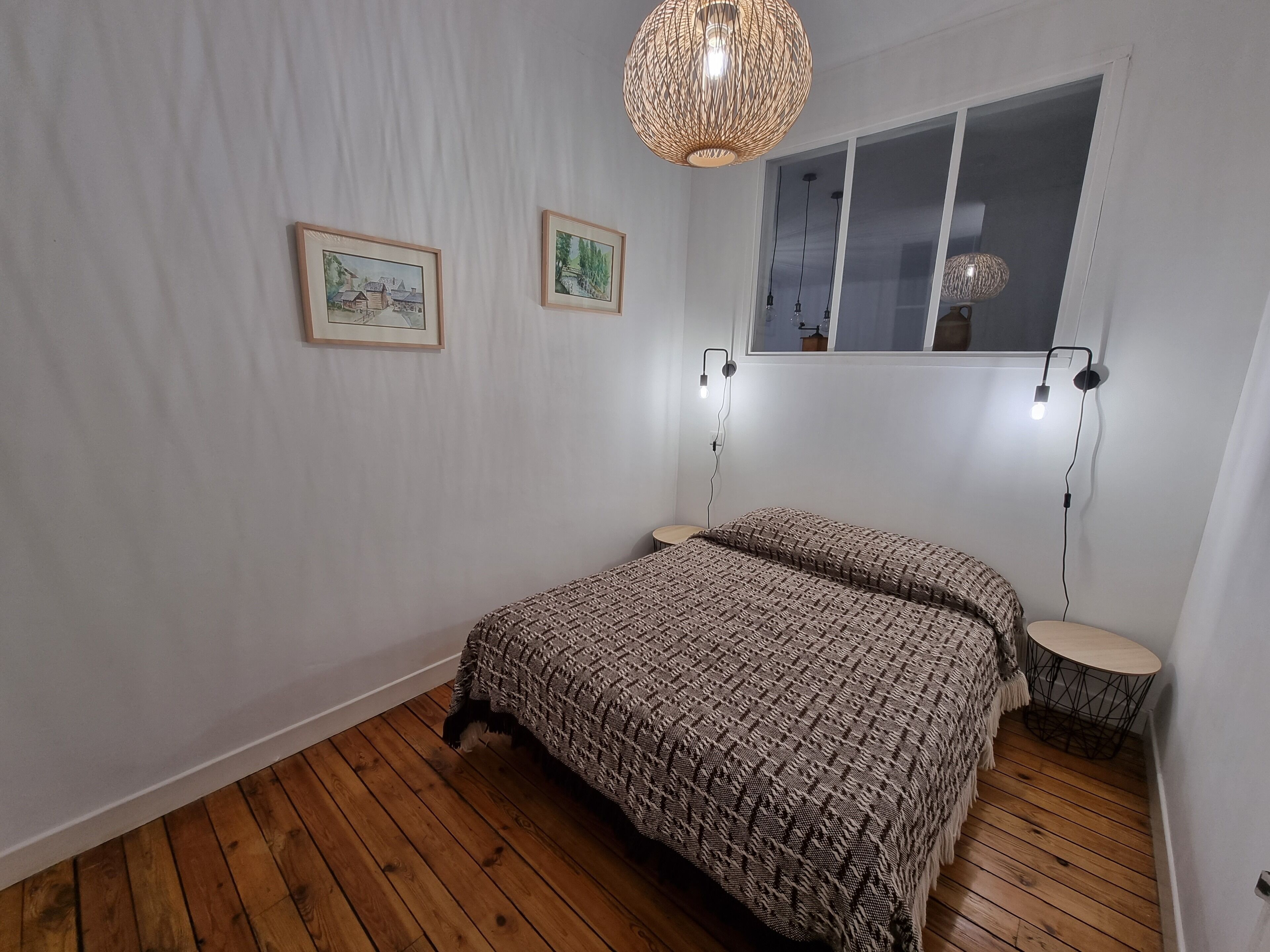 A bedroom with a wooden floor, a bed with patterned bedding, two wall-mounted lamps, and two framed pictures.