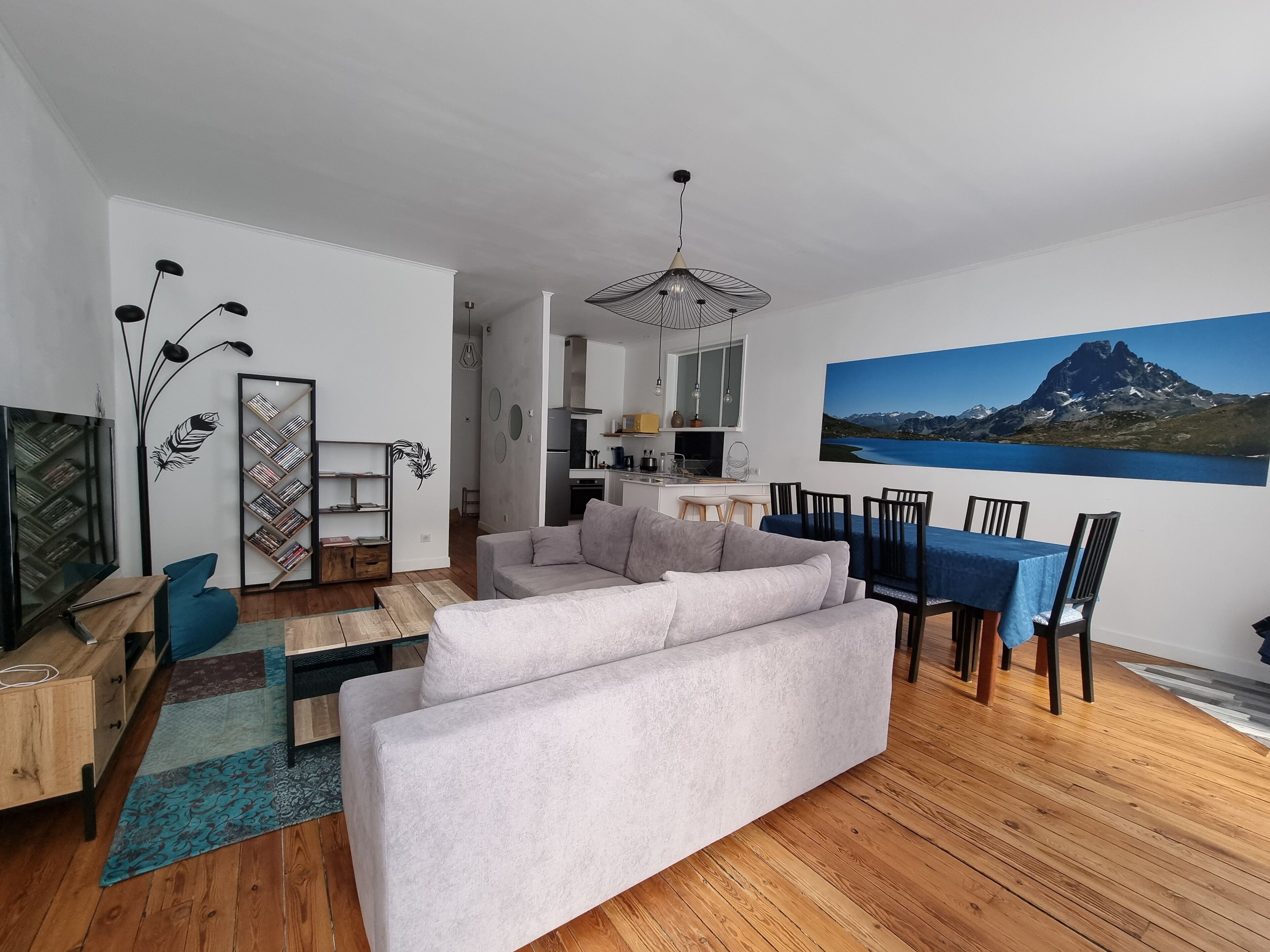 Un salon moderne comprenant un canapé, une table à manger avec des chaises, une bibliothèque et un grand tableau représentant un paysage de montagne.