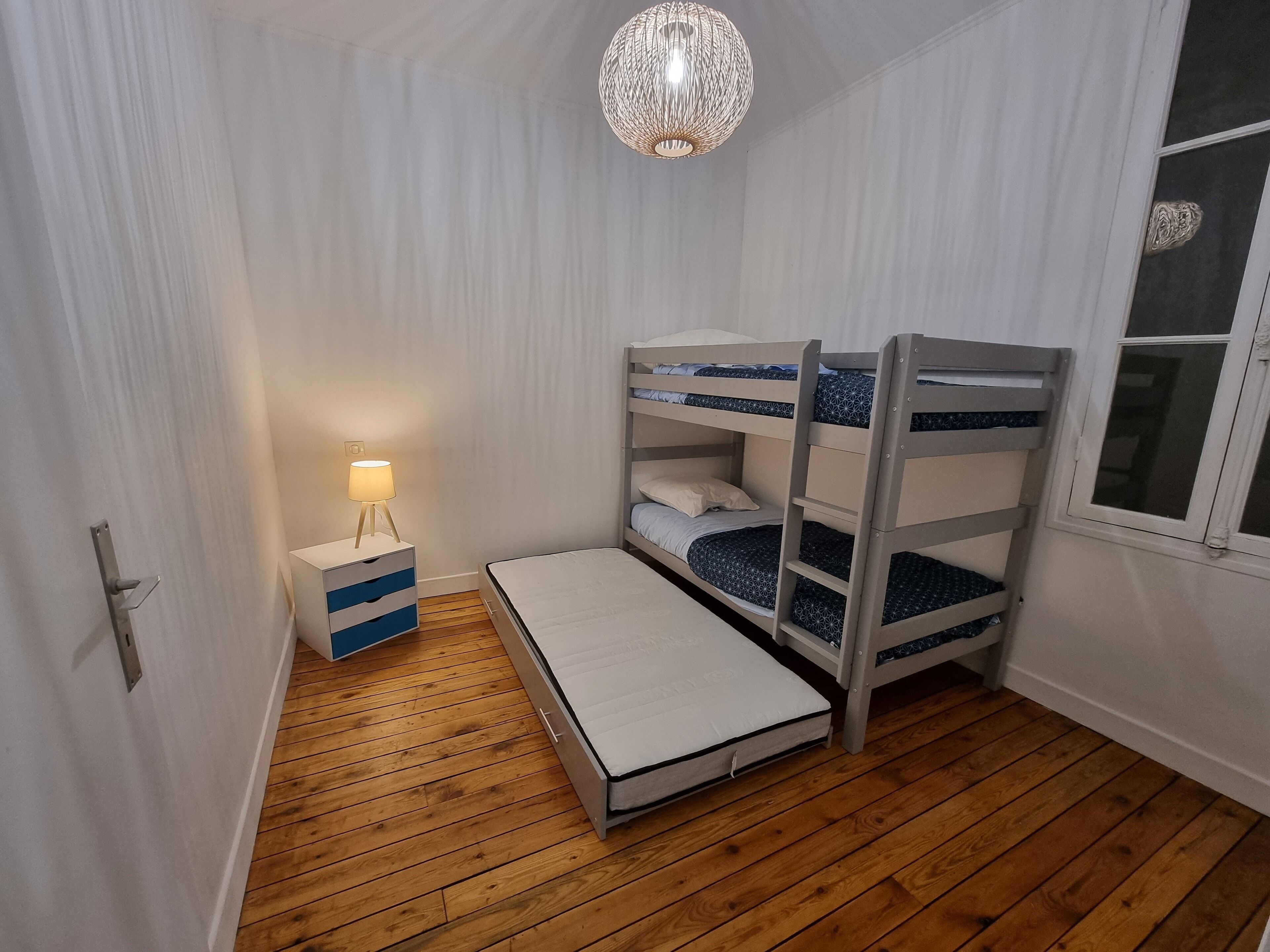 A bunk bed room with a bedside table and lamp, wooden flooring, and a window with a view.