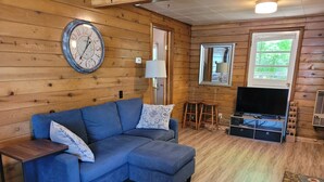 A cozy room with a blue sectional sofa, a wooden wall clock, a TV, and a window with a view of greenery.