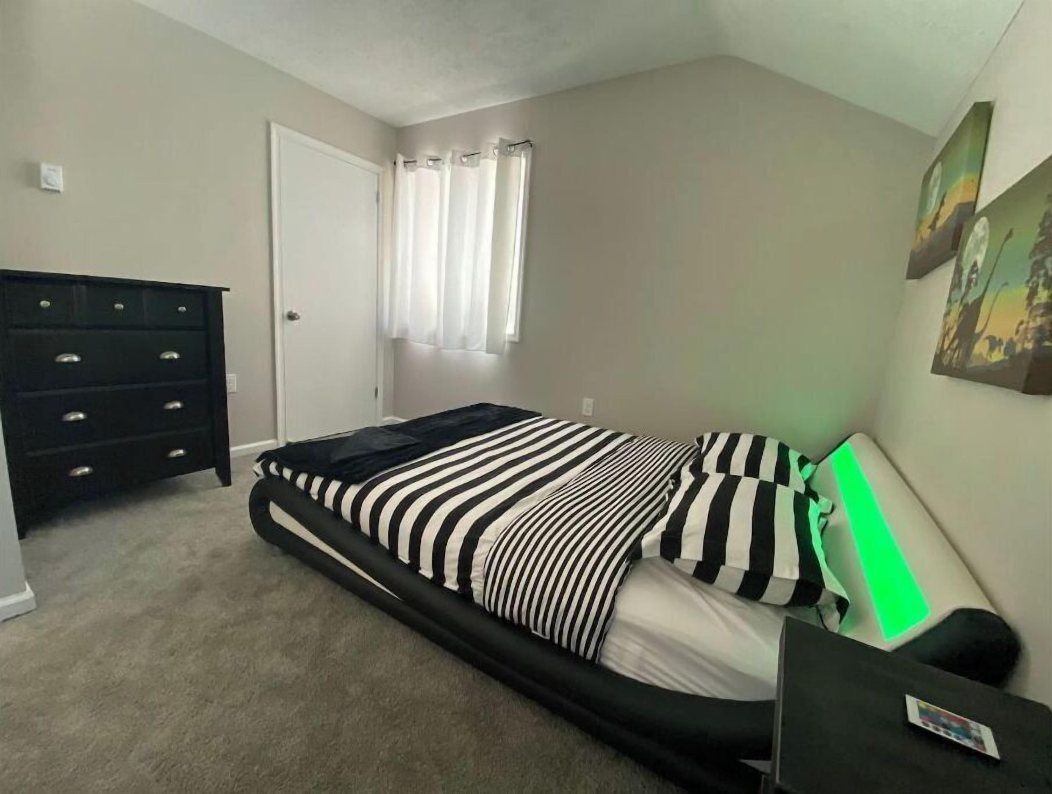 Una camera da letto con un letto, una cassettiera, una finestra con tende e un quadro appeso al muro.