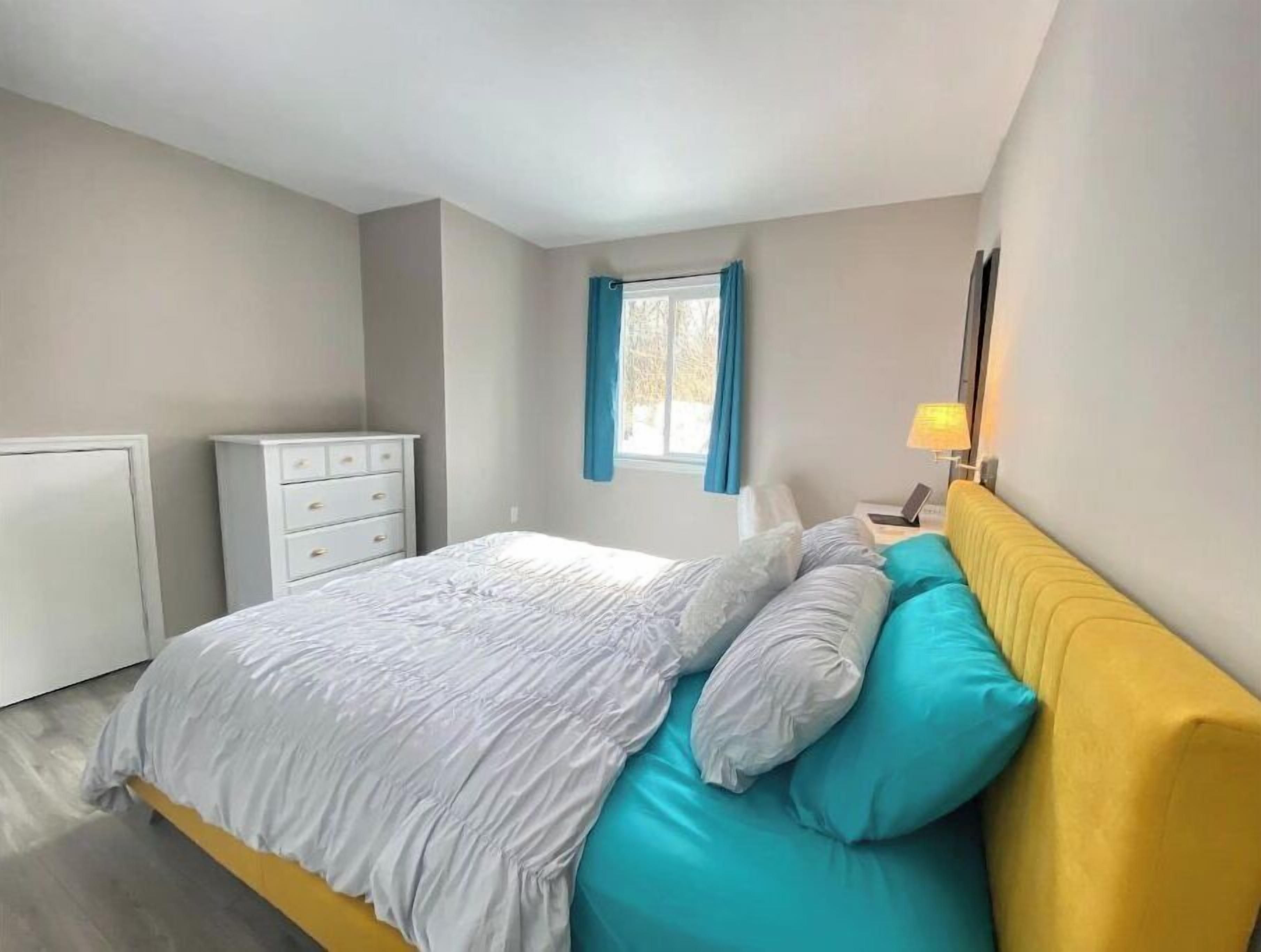 Un dormitorio con una cama, una mesita de noche con lámpara, una ventana con cortinas y un aparador blanco.
