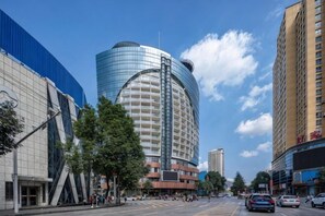 A modern hotel with a curved glass facade, a prominent entrance, and a parking lot with cars. - Ausotel Hotel (Jinsha)