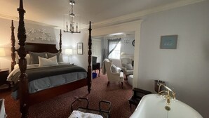 Una cama con dosel, un espacio de estar con una silla y una mesa, una bañera y una puerta que da a otra habitación.