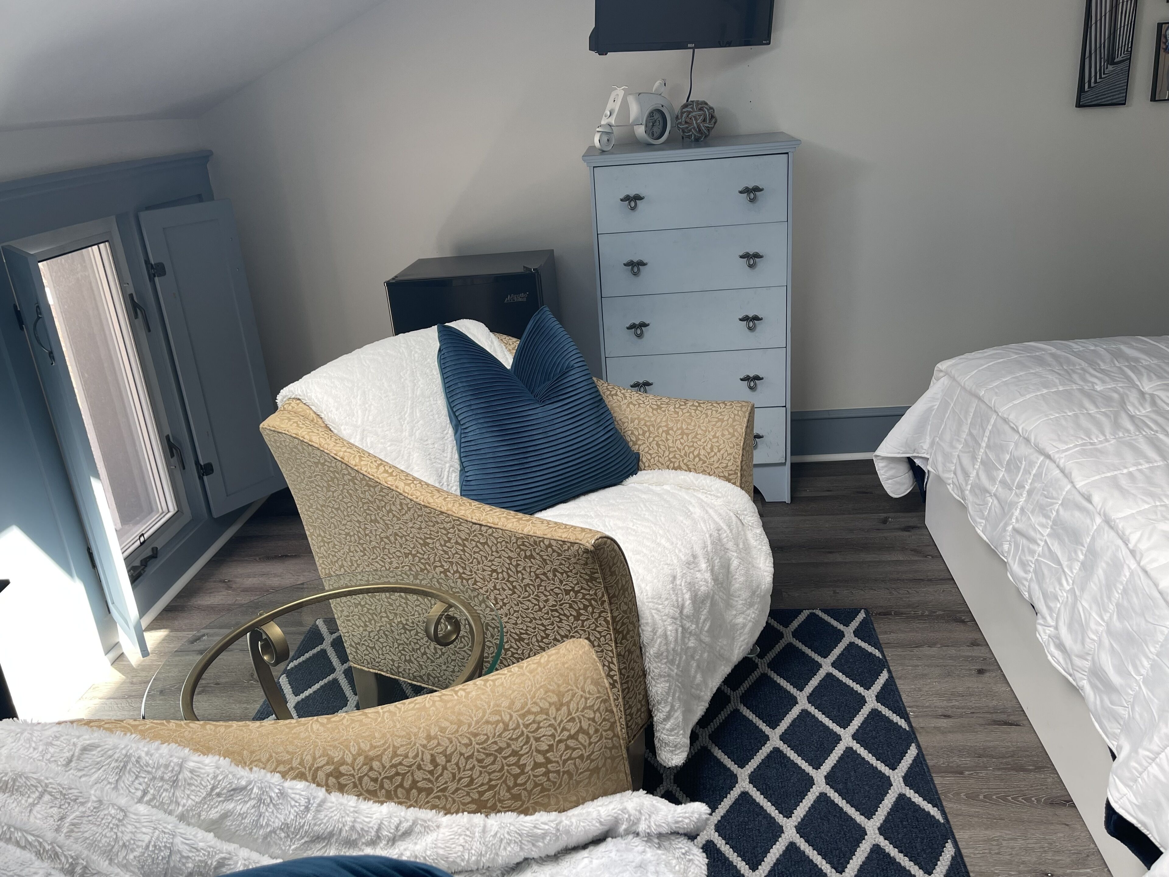 Ein Schlafzimmer mit einem Bett, einem Sessel mit Decke, einem Schminktisch und einem an der Wand befestigten Fernseher.