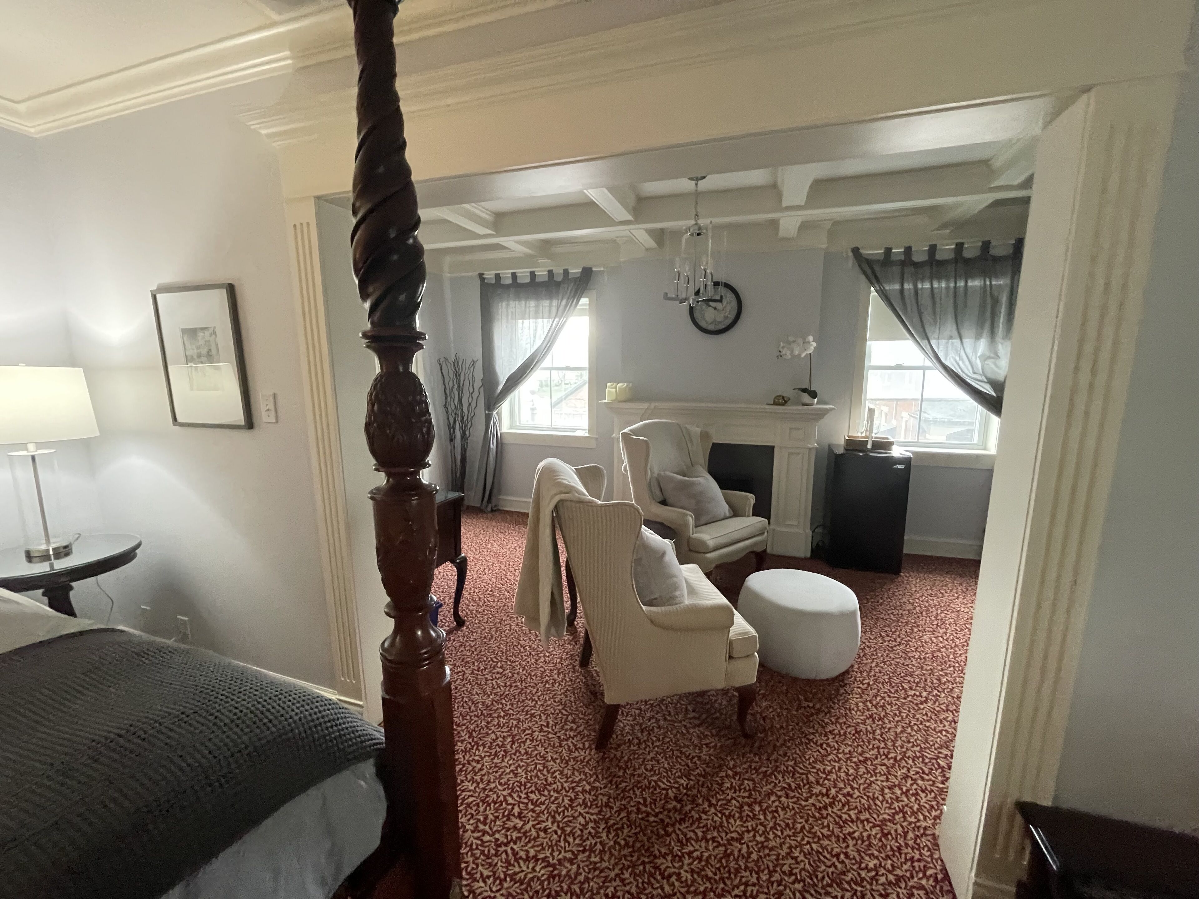 A bedroom with a four-poster bed, a sitting area with chairs and a table, a fireplace, and a large window with curtains.