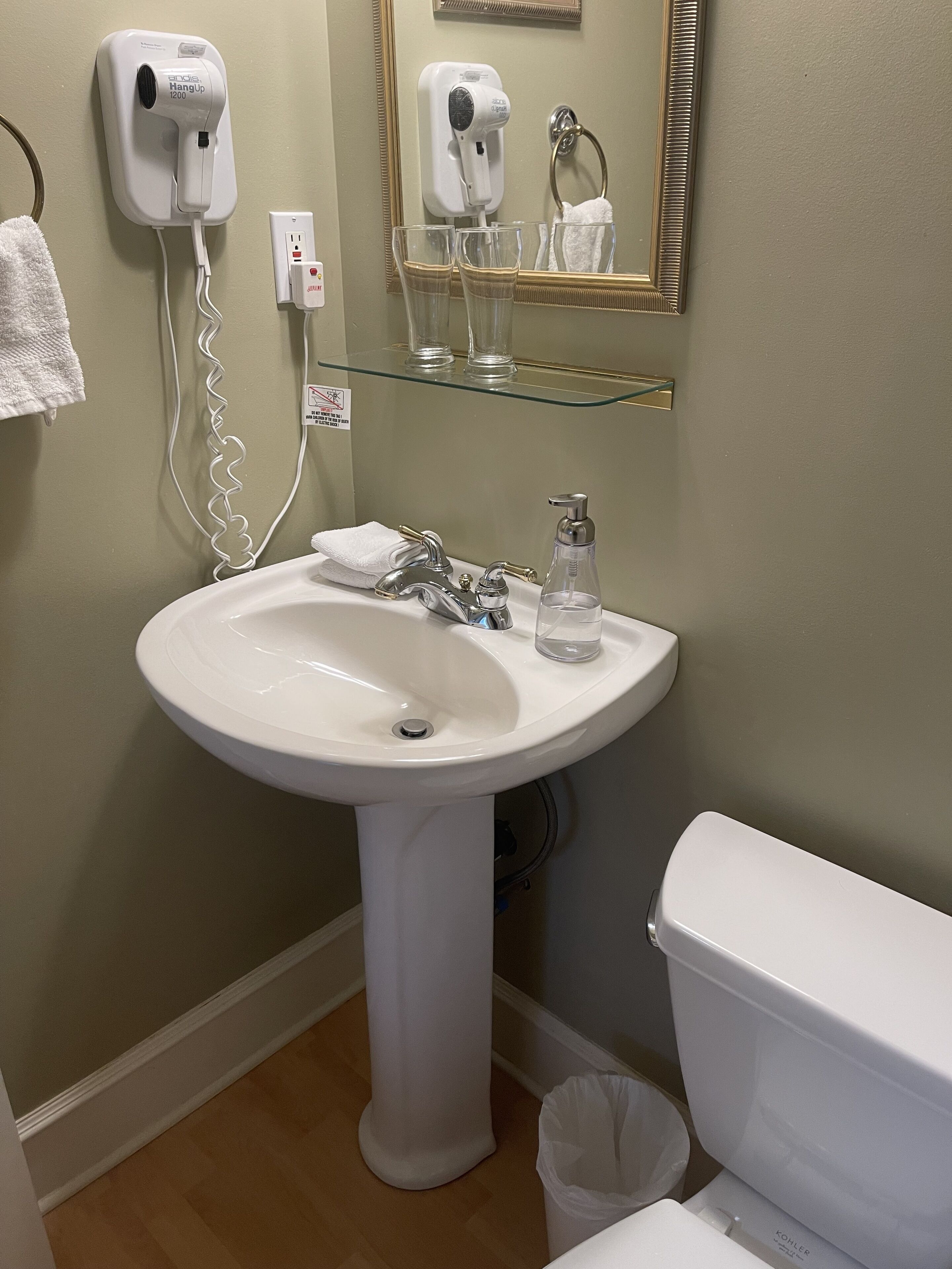 A bathroom with a white pedestal sink, a Kohler toilet, a hair dryer, and a glass shelf with a towel and a glass.