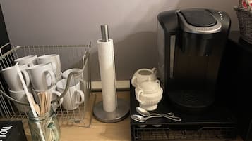 En köksbänk med en kaffebryggare, en korg med köksredskap, en burk och ett ställ för kaffemuggar.