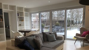 A modern living room with a grey sectional sofa, a white armchair, and a dining table with chairs. There is a bookshelf and a large sliding glass door leading to an outdoor patio area. - Luxurious country villa nestled in a horse farm on the mountain with great views (Uttenhofen)