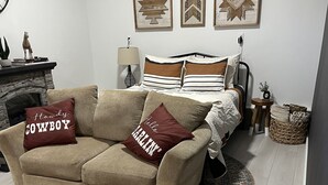 Een gezellige woonkamer met een beige bank, twee decoratieve kussens, een kamerkleed met een patroon, een bed met gestreept beddengoed en een klein bijzettafeltje met een plantje.