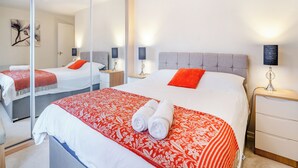 A hotel room with a large mirror, a bed with a red and white patterned bedspread, two bedside tables with lamps, and a wall-mounted light fixture.