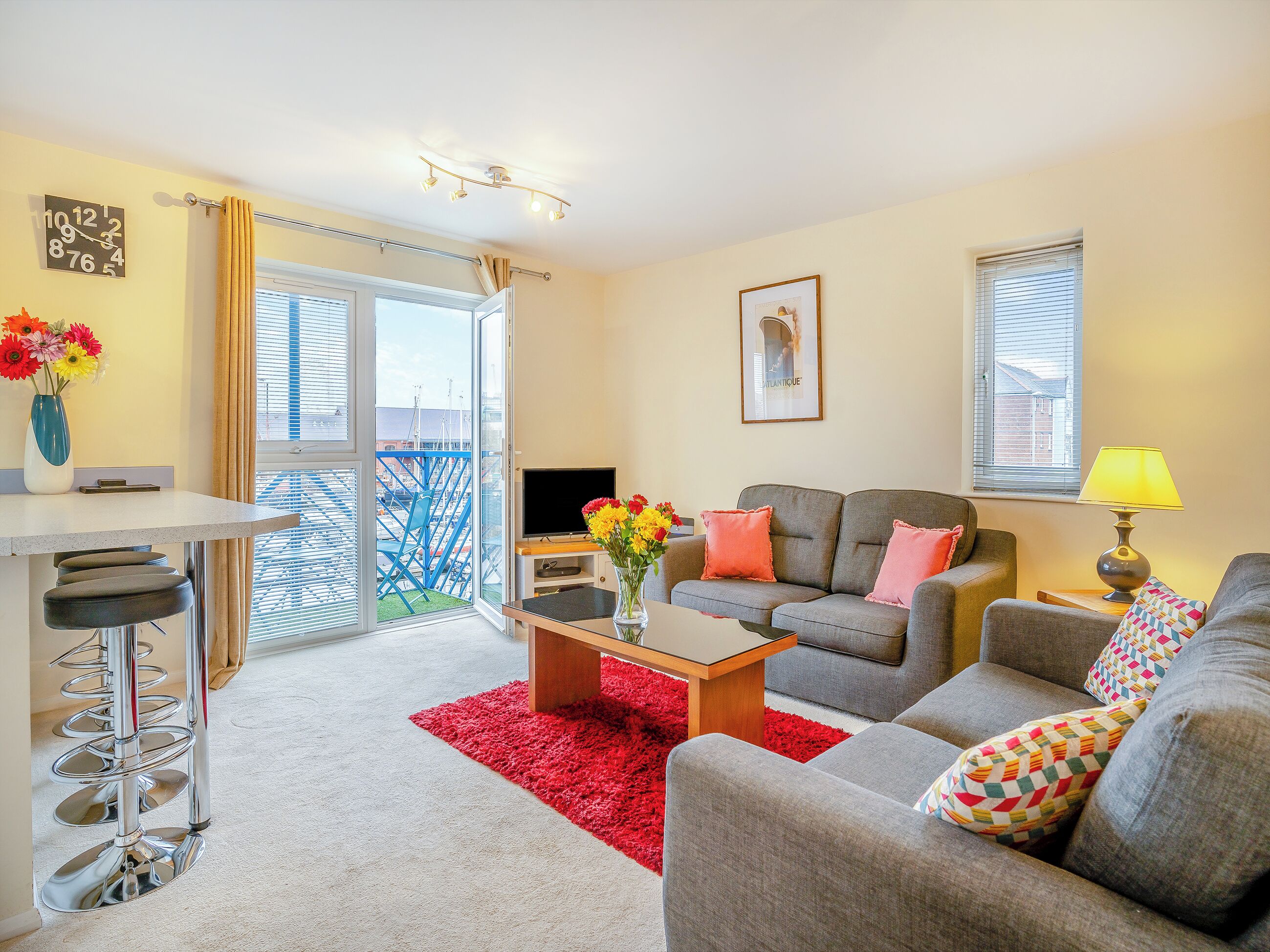 A living room with a grey sofa, a red rug, and a TV. There is a balcony with blue chairs and a railing.