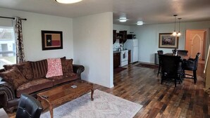 Een woonkamer met een bruine bank, een houten salontafel en een eethoek met een donkerhouten tafel en stoelen. In de achtergrond is een keuken met witte apparatuur zichtbaar.