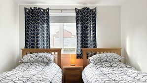 A bedroom with two single beds, a nightstand, a lamp, and a window with curtains. - Academy Street Cottage (Tain)